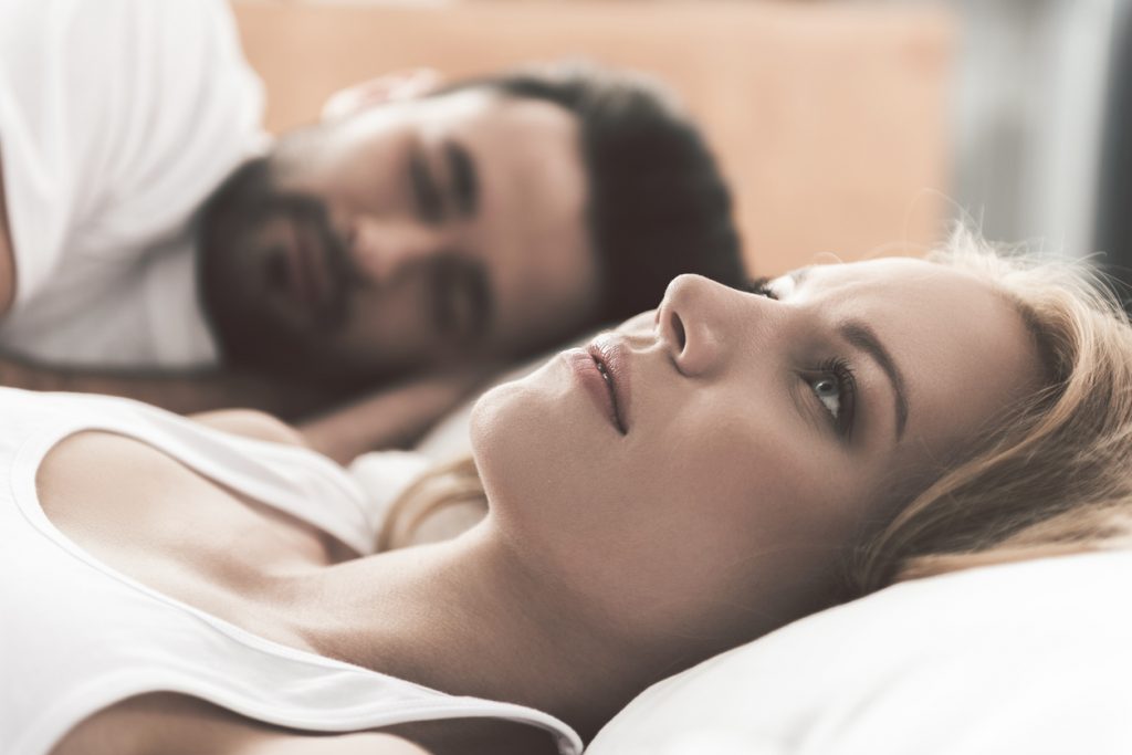 Nervous young woman is lying on bed and thinking seriously. Her husband is sleeping on background